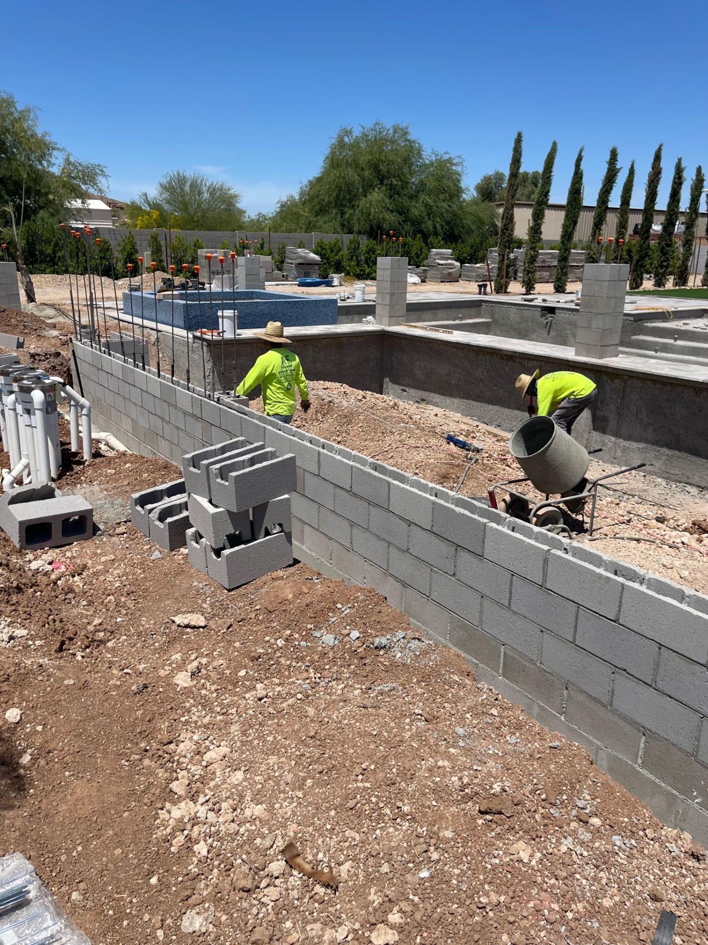 Aurora Estate — masonry crew building the retaining wall beside the empty pool shell with the raised mosaic-tile spa and cypress trees in the background