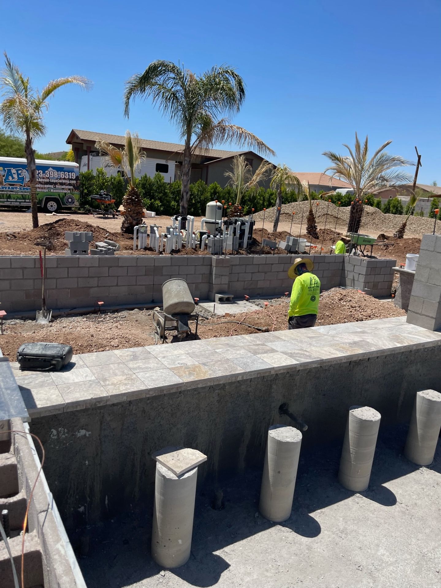 Aurora Estate — view from inside the empty pool shell toward the equipment wall, masonry runs, and palms as structural systems converge
